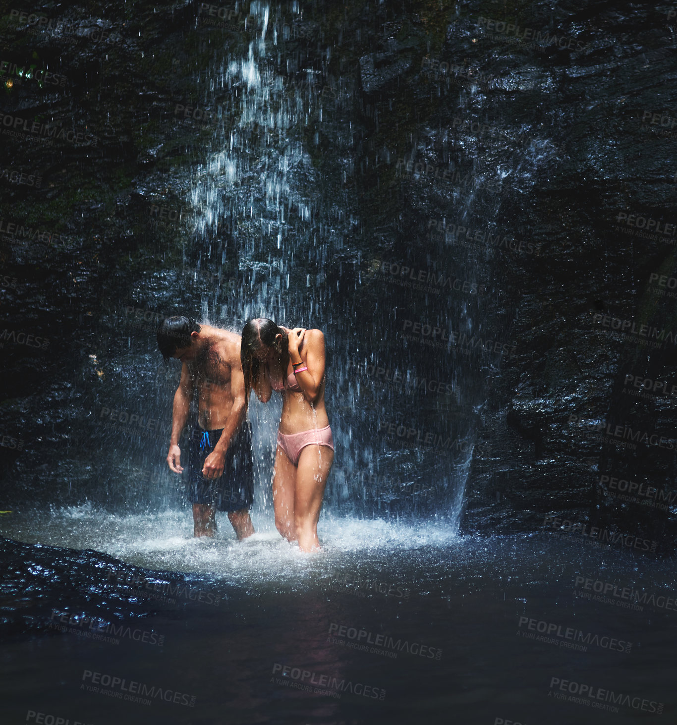 Buy stock photo Shot of two young people standing under a waterfall and enjoying every moment of it while looking down