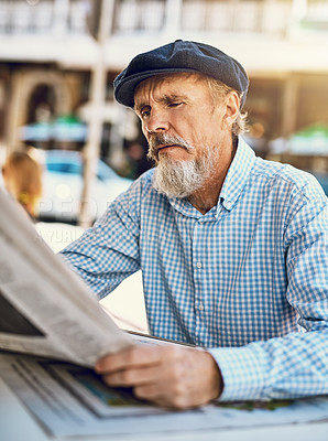 Buy stock photo Shot of a serious senior man reading the newspaper while sitting at a sidewalk cafe