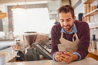 Buy stock photo Shot of a happy business owner using his smartphone while leaning on the counter in his coffee shop