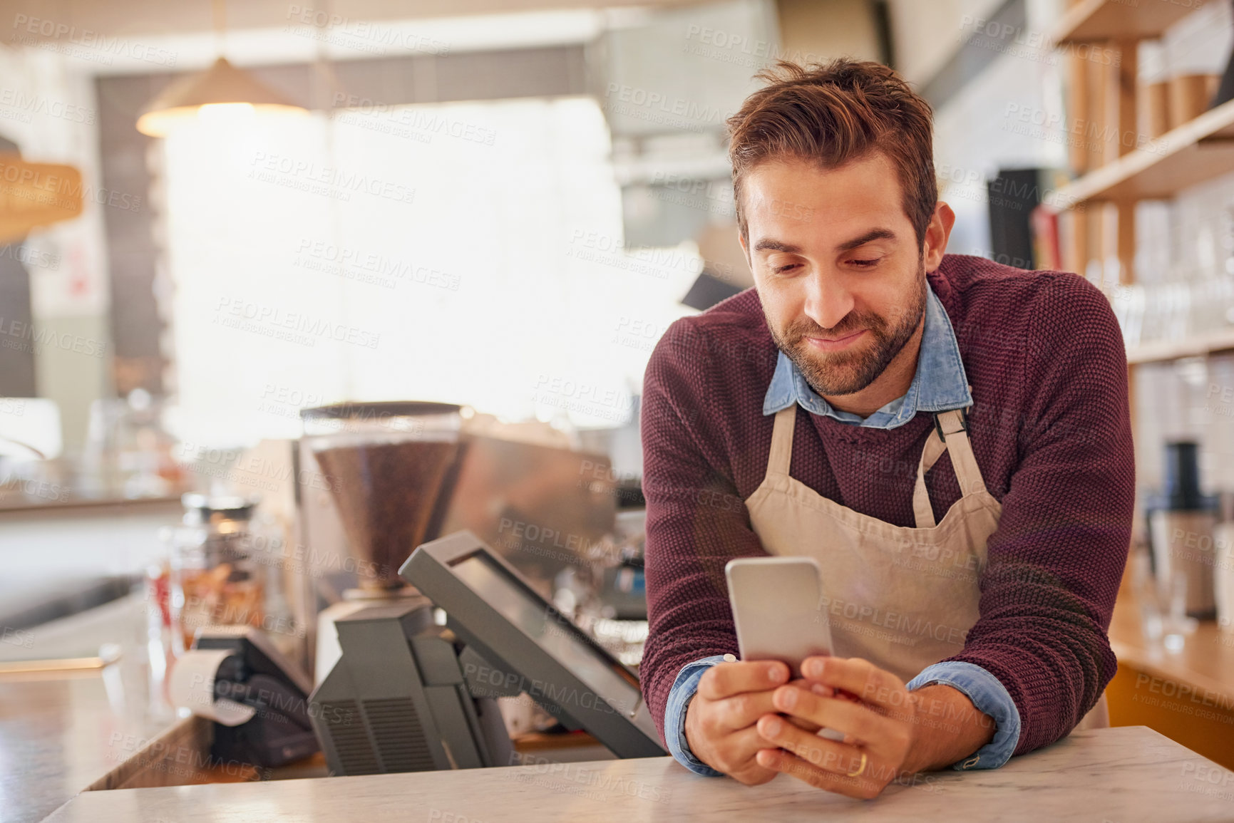 Buy stock photo Shot of a happy business owner using his smartphone while leaning on the counter in his coffee shop