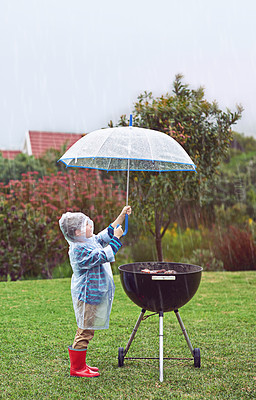 Buy stock photo Full length shot of a young boy standing in front of an outdoor grill in the rain