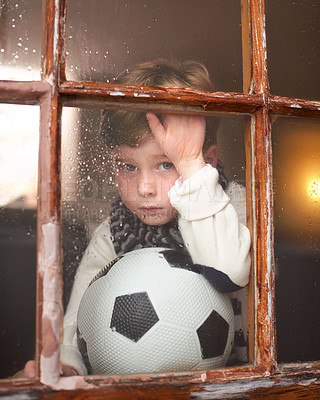 Buy stock photo Shot of a sad little boy holding a soccer ball while watching the rain through a window