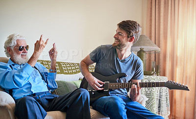 Buy stock photo Shot of a happy young man playing the electric guitar while his elderly grandfather makes a rock on gesture on the couch