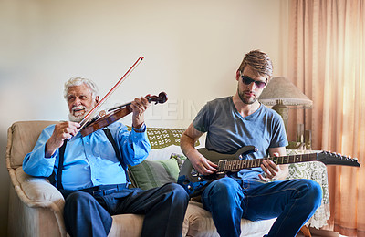Buy stock photo Shot of a young man playing the electric guitar while his elderly grandfather plays the violin on the couch