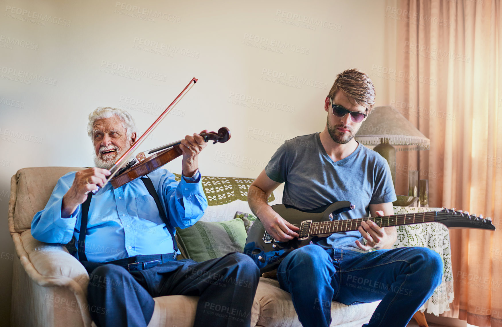 Buy stock photo Shot of a young man playing the electric guitar while his elderly grandfather plays the violin on the couch