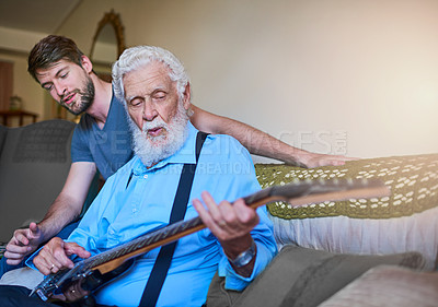 Buy stock photo Shot of a happy young man teaching his elderly grandfather to play the electric guitar on the couch at home