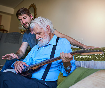 Buy stock photo Shot of a happy young man teaching his elderly grandfather to play the electric guitar on the couch at home