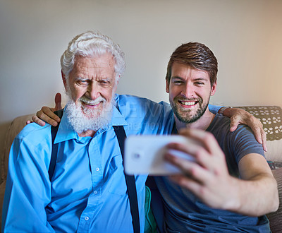 Buy stock photo Shot of a happy young man and his elderly grandfather posing for a selfie together on the couch at home