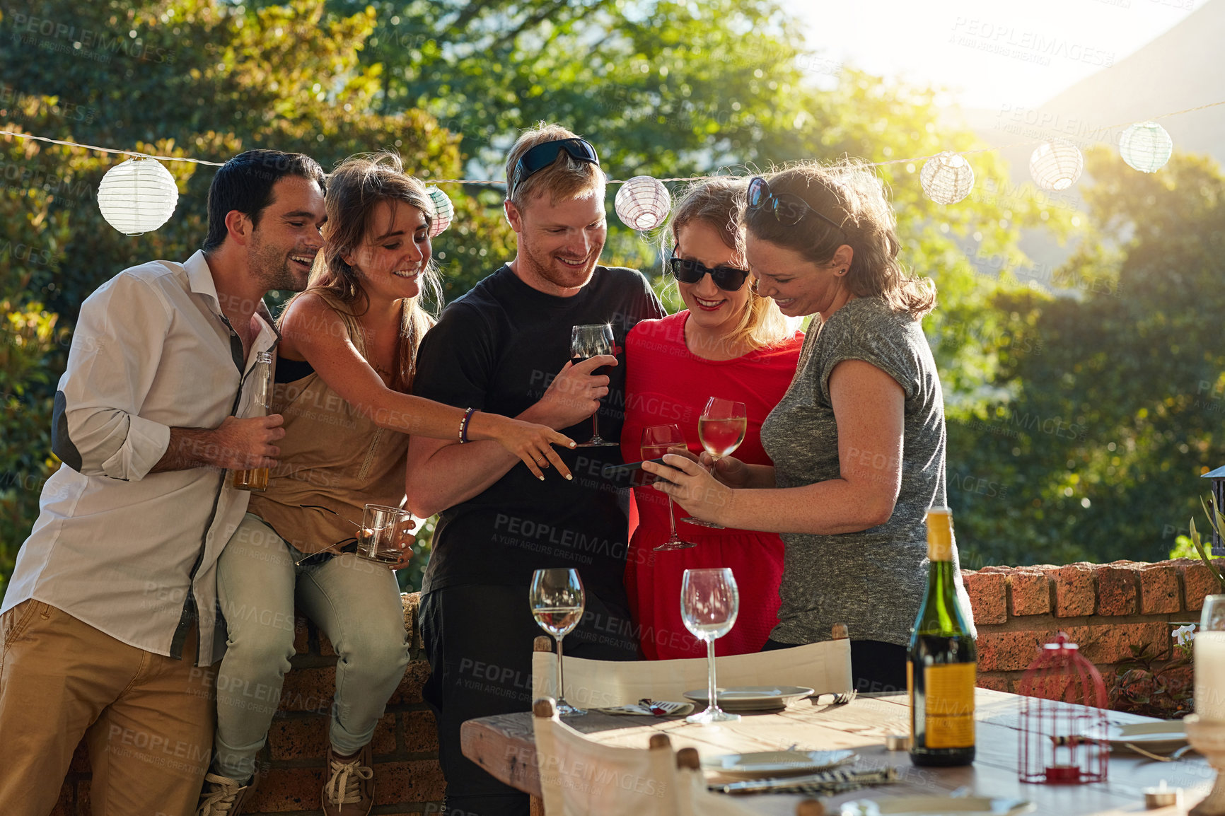 Buy stock photo Friends, phone and drinks in backyard, party and happy with video, meme or post on social media. People, men and woman with wine, alcohol and smartphone at event on mobile app in sunshine on holiday