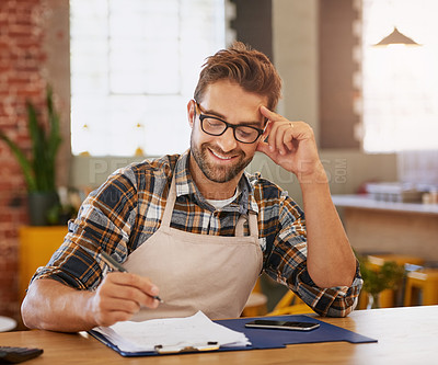 Buy stock photo Shot of a happy young business owner doing admin at a table in his coffee shop