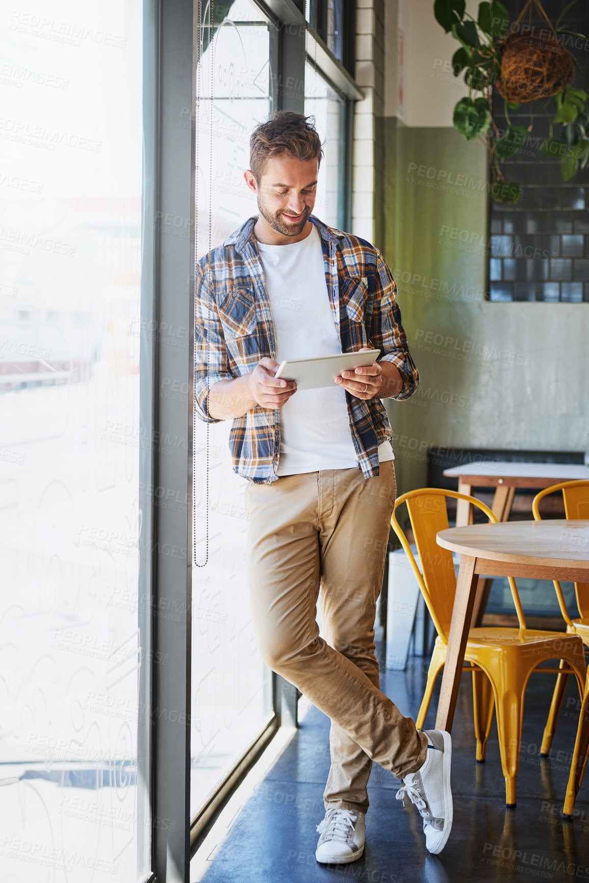Buy stock photo Shot of a relaxed young man using his tablet while leaning against a window in his favorite coffee shop