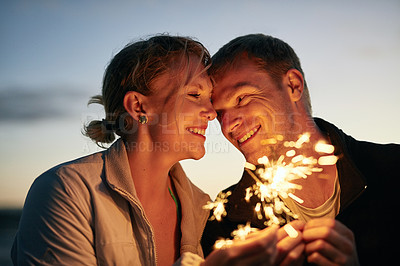 Buy stock photo Shot of a happy young couple playing with sparklers on the beach at sunset