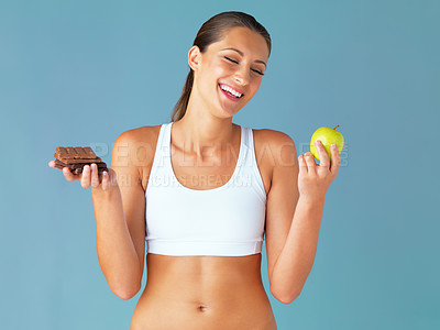 Buy stock photo Studio shot of a fit young woman deciding whether to eat a chocolate or an apple against a blue background