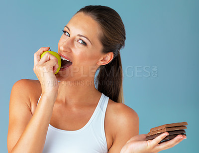 Buy stock photo Studio shot of a young woman holding a chocolate while taking a bite of an apple against a blue background
