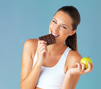 Buy stock photo Studio shot of a fit young woman holding an apple while taking a bite of chocolate against a blue background