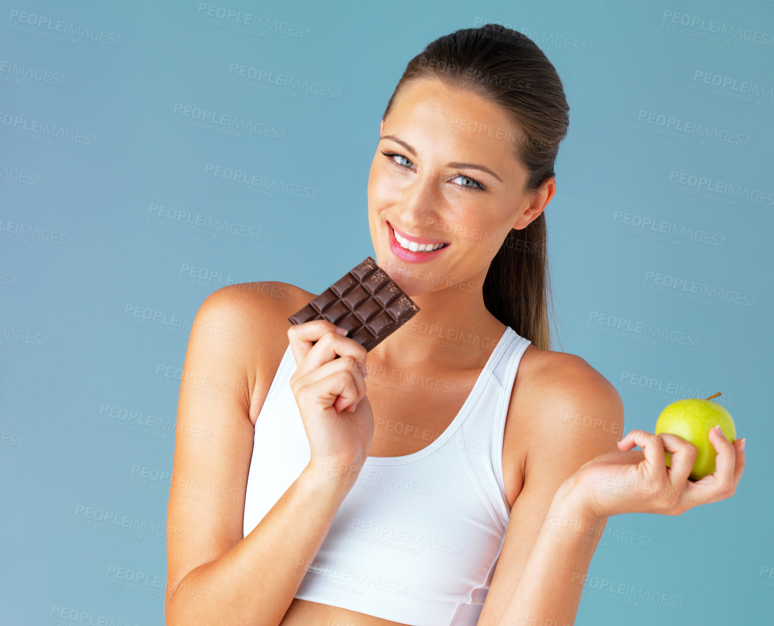 Buy stock photo Studio shot of a fit young woman holding an apple and a chocolate against a blue background
