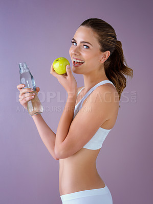 Buy stock photo Studio shot of a healthy young woman holding a glass water bottle and an apple against a purple background