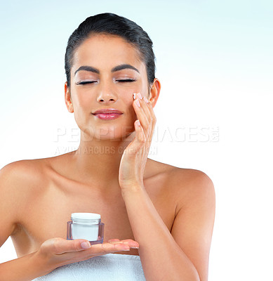 Buy stock photo Studio shot of a beautiful young woman applying moisturizer onto her face with her eyes closed against a blue background
