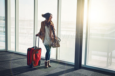 Buy stock photo Shot of a young woman standing in an airport with her luggage and staring outside
