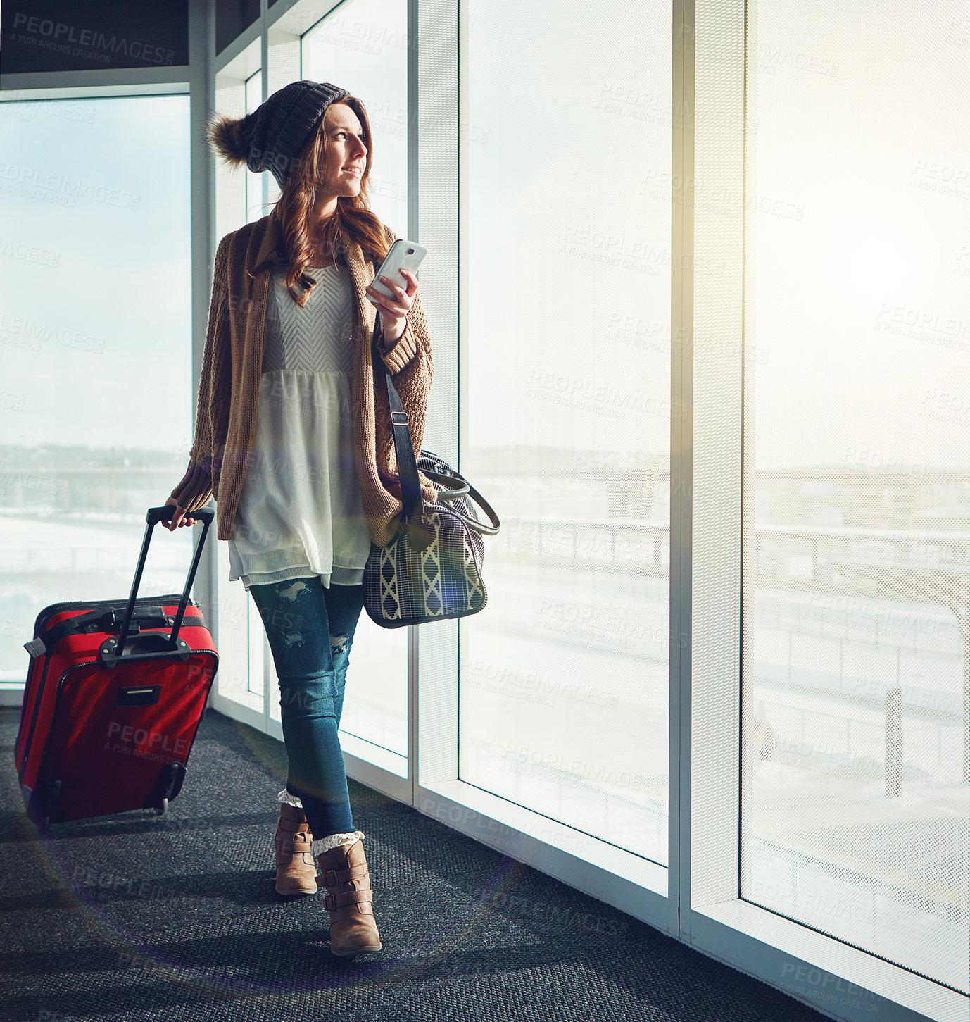 Buy stock photo Shot of a young woman standing at an airport with her luggage staring outside while holding her cellphone