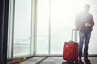 Buy stock photo Shot of a young man standing in an airport with his luggage while looking at his cellphone and texting