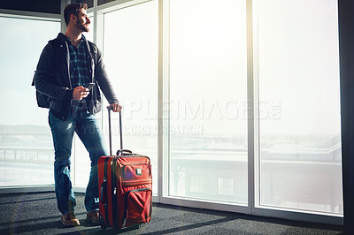 Buy stock photo Shot of a young man standing in an airport with luggage and looking outside while holding his cellphone