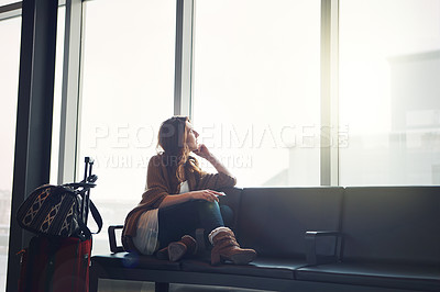 Buy stock photo Shot of a young woman sitting inside of an airport with her luggage looking outside with her cellphone in her hand