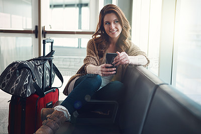 Buy stock photo Shot of a young woman sitting in an airport with her luggage and holding her cellphone while looking outside