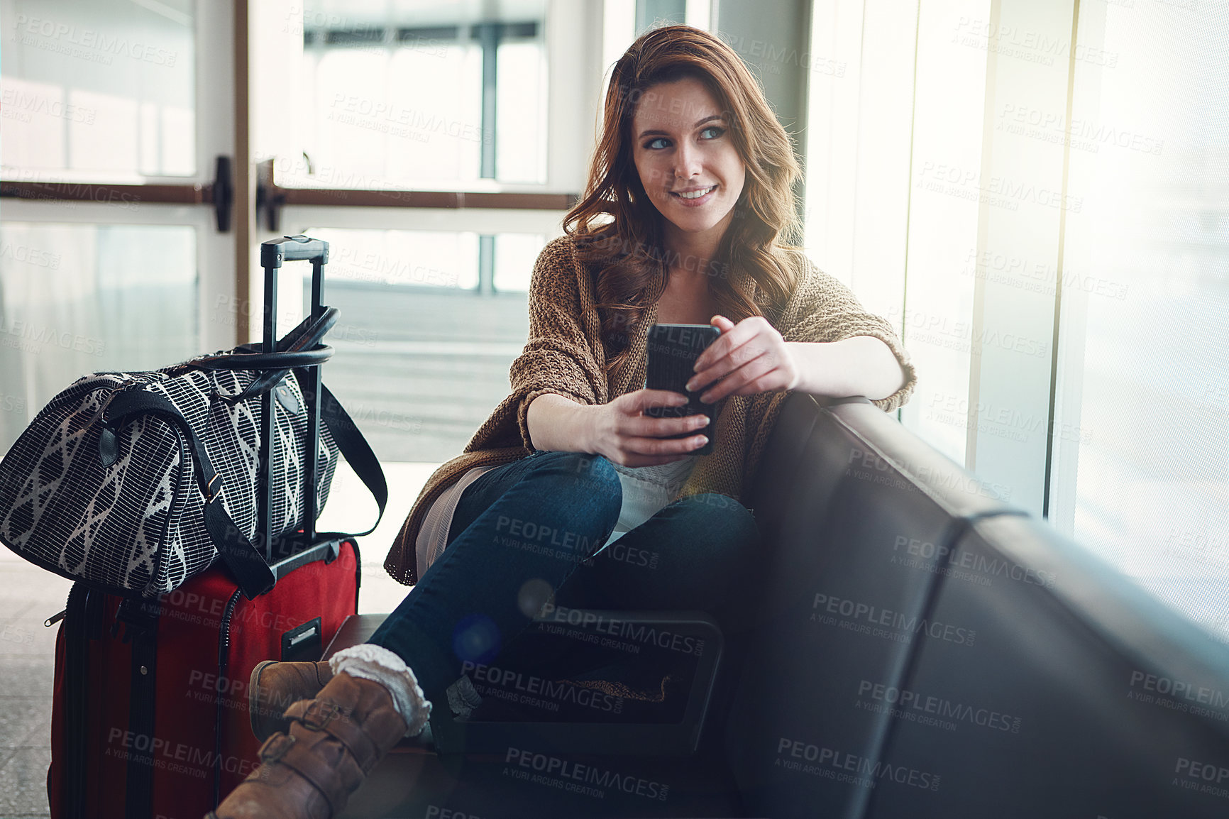 Buy stock photo Shot of a young woman sitting in an airport with her luggage and holding her cellphone while looking outside