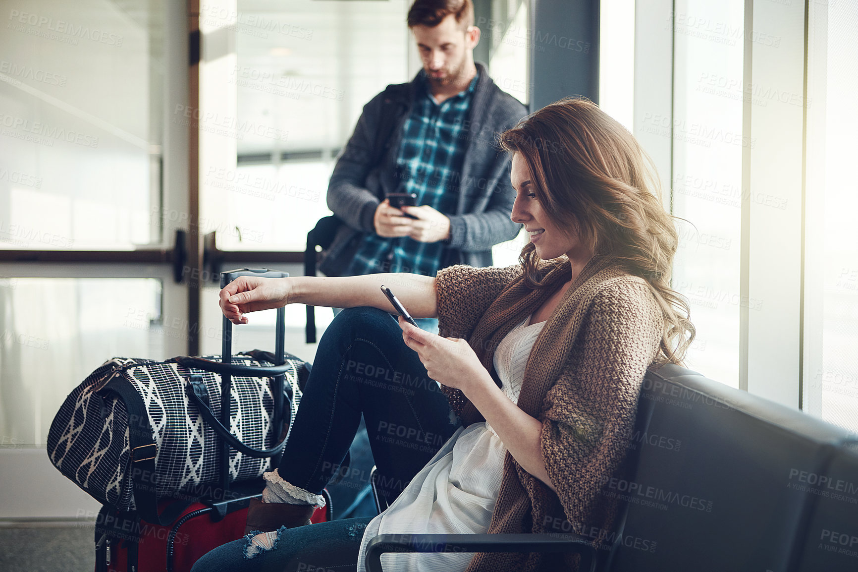 Buy stock photo Shot of a young couple sitting in an airport with their luggage while making use of their cellphones