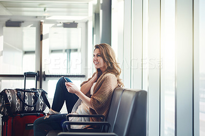 Buy stock photo Shot of a young woman sitting in an airport with her luggage while holding her phone and looking at something
