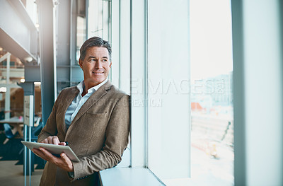 Buy stock photo Shot of a mature businessman standing in the office and making notes on his tablet while looking through a window