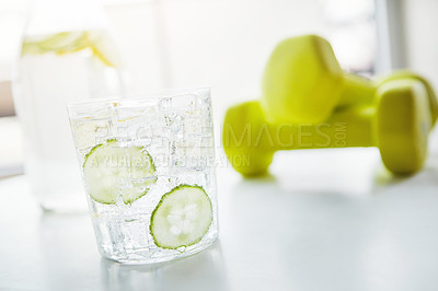 Buy stock photo Glass, water and cucumber with dumbbell on table for detox, nutrition and weight loss in house. Fitness, beverage and liquid with ice in container for vitamin C, antioxidants and organic hydration