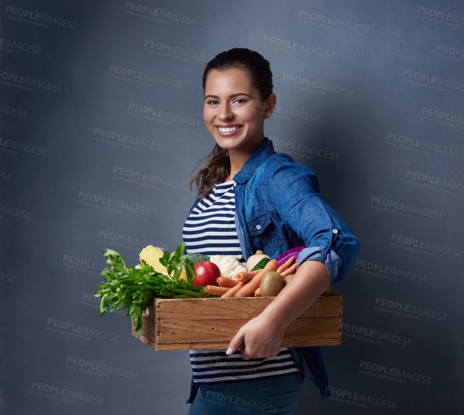 Buy stock photo Studio shot of a beautiful young woman holding a wooden crate full of vegetables and fruit against a blue background