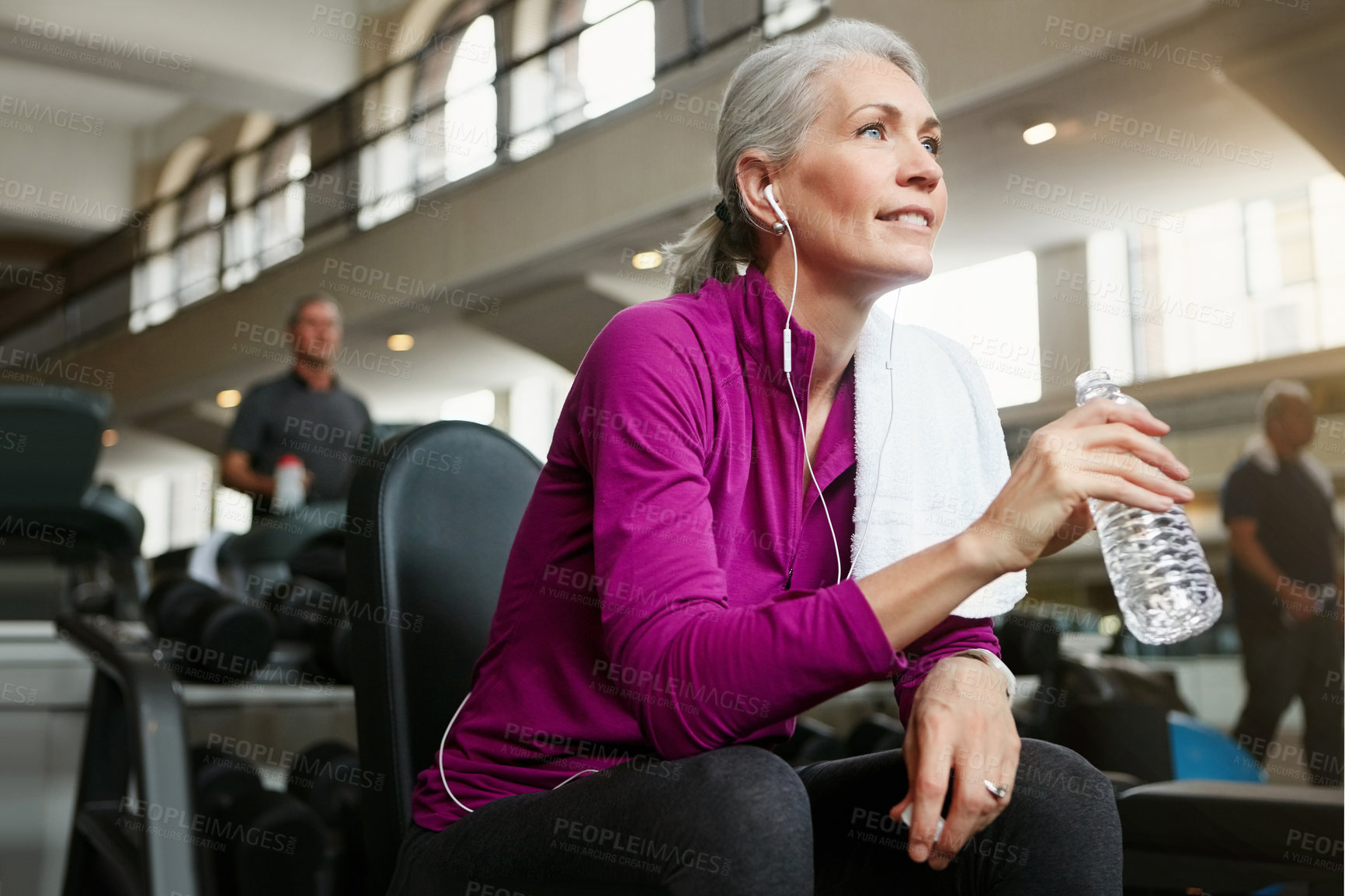 Buy stock photo Mature woman, break and drinking with water, earphones or music on machine for workout or exercise at gym. Female person with smile for rest, mineral beverage or thirst after training at health club