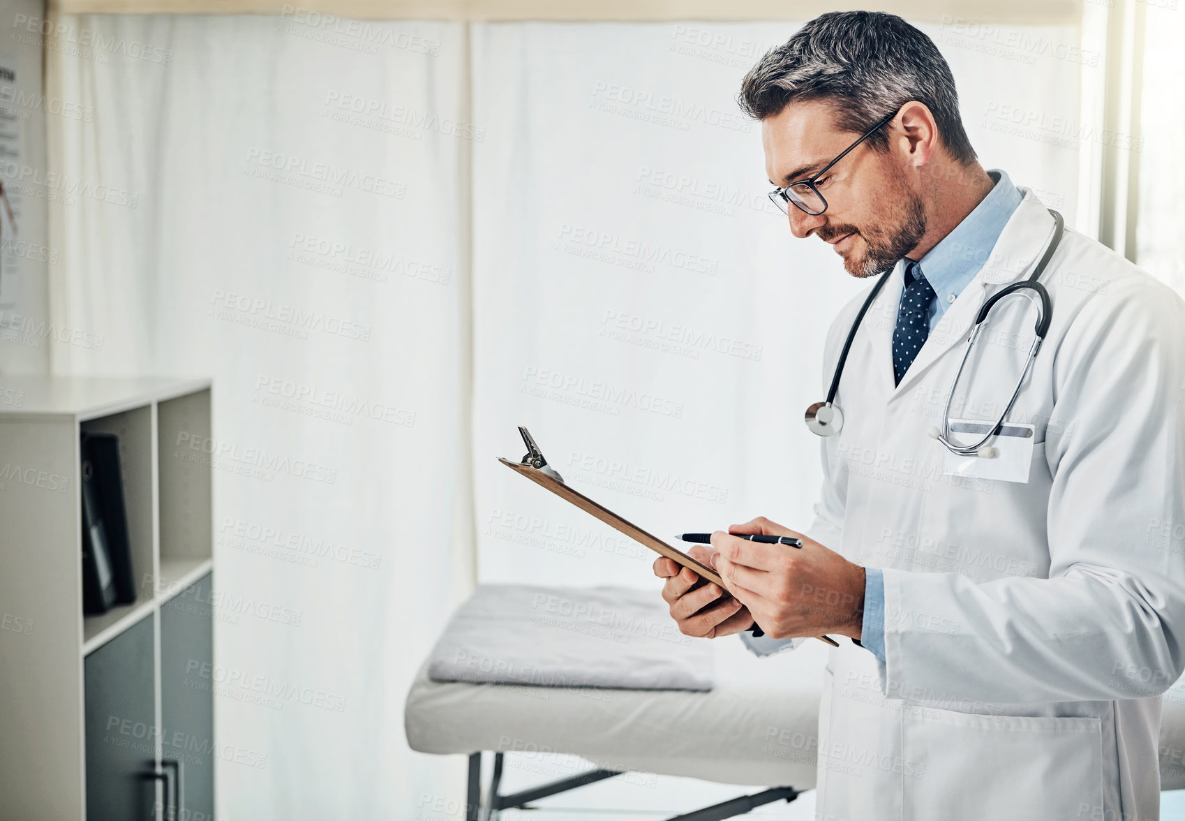 Buy stock photo Shot of a focussed doctor reading a patient file while standing in his consultation room