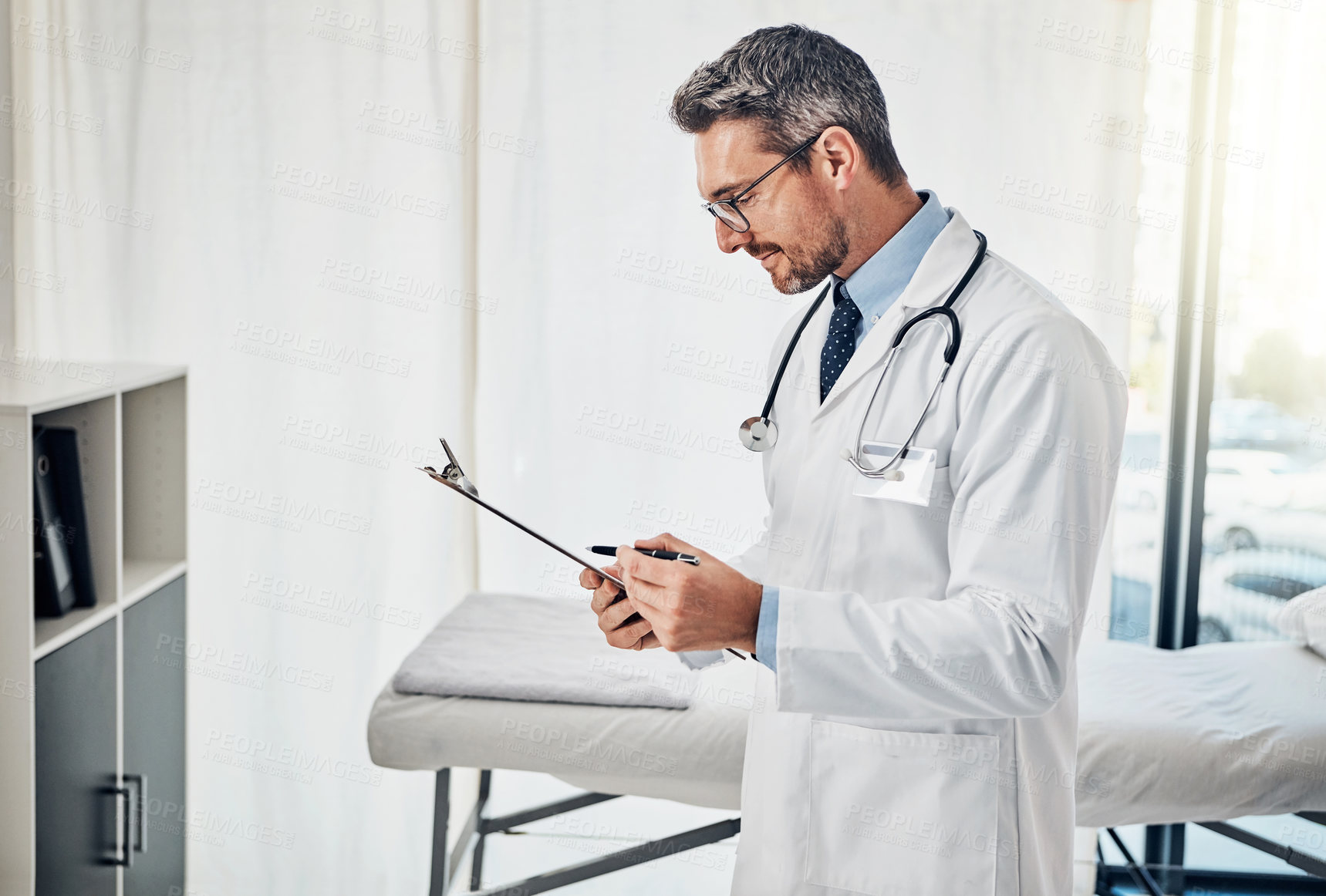 Buy stock photo Shot of a focussed doctor reading a patient file while standing in his consultation room