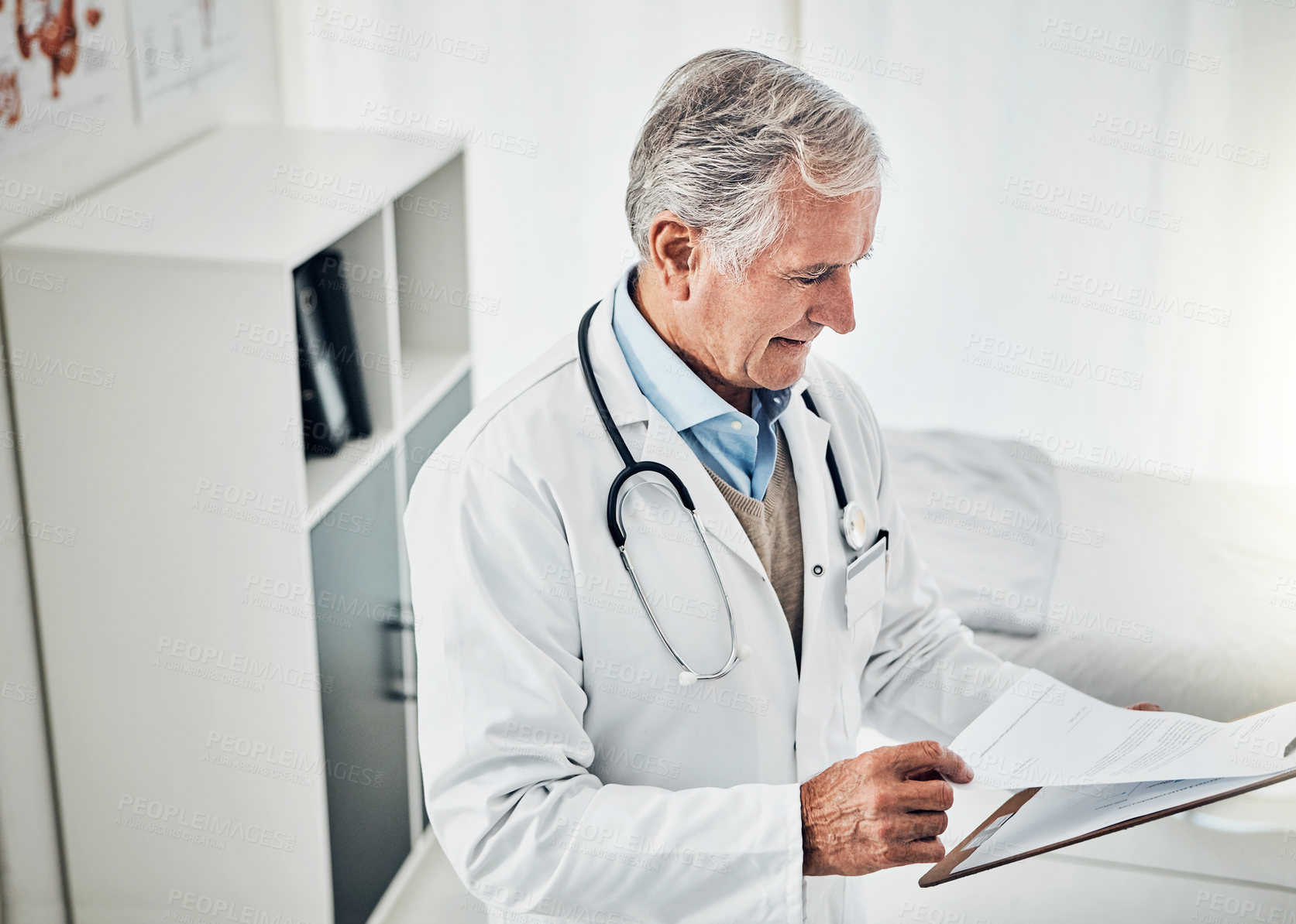 Buy stock photo Shot of a mature doctor reading a patient file while standing in his consultation room