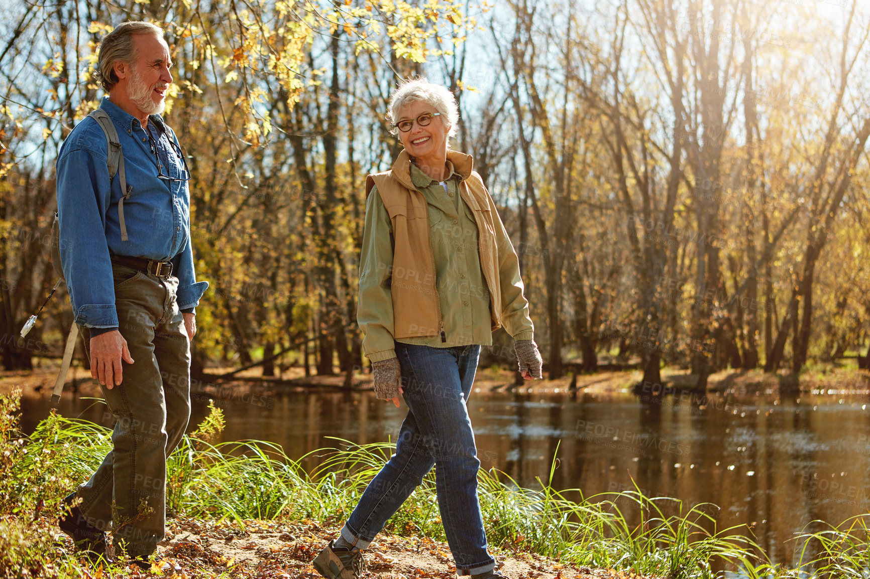 Buy stock photo Old couple, laugh and hiking in forest with talk, funny story and happy on adventure in countryside. Elderly man, woman and smile for bonding, care and lake with trekking in woods in retirement