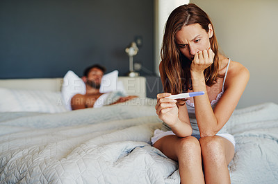 Buy stock photo Shot of a woman looking worried while holding a pregnancy test with her partner lying in the background