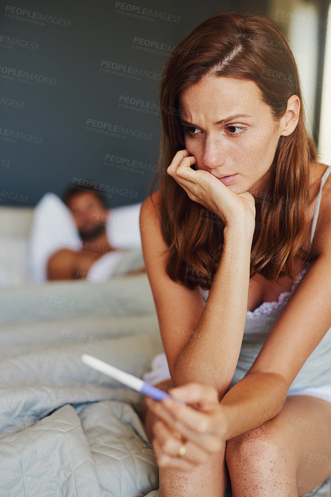 Buy stock photo Shot of a woman looking worried while holding a pregnancy test with her partner lying in the background