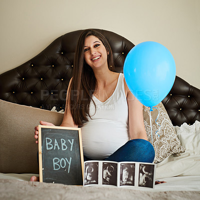 Buy stock photo Portrait of a happy pregnant woman posing on her bed with an ultrasound picture, chalkboard and blue balloon