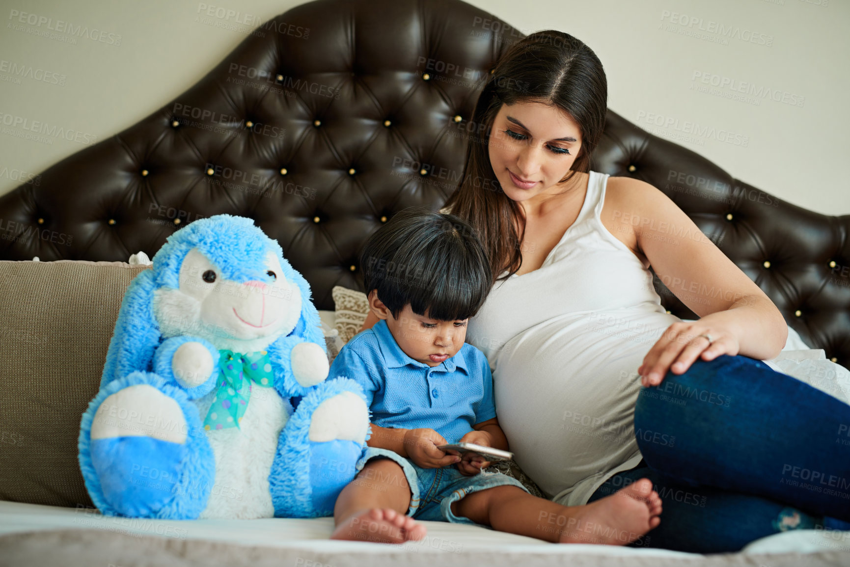 Buy stock photo Shot of a pregnant woman looking at her little boy while he plays with a smartphone on her bed