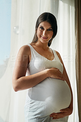 Buy stock photo Portrait of a pregnant woman cradling her belly while standing in front of a window at home