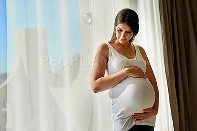Buy stock photo Shot of a pregnant woman cradling her belly while standing in front of a window at home