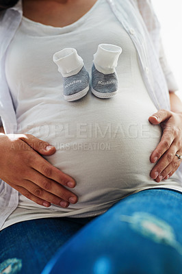 Buy stock photo Portrait of an unidentifiable pregnant woman sitting in a chair at home with a pair of baby shoes on her belly