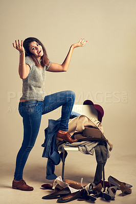 Buy stock photo Studio shot of a young woman choosing clothing piled on a chair against a brown background
