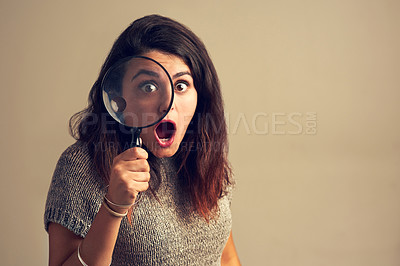 Buy stock photo Studio portrait of a young woman looking shocked while using a magnifying glass against a brown background