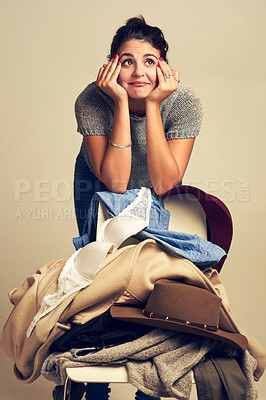 Buy stock photo Studio shot of a thoughtful young woman choosing clothing piled on a chair against a brown background