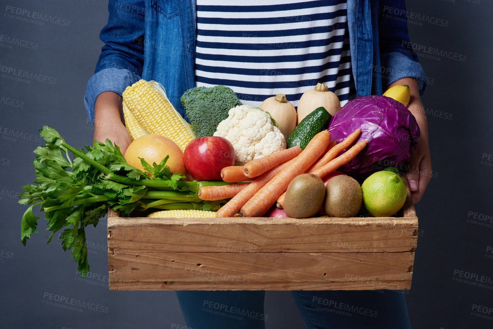 Buy stock photo Studio shot of an unrecognizable woman holding a wooden crate full of fruit and vegetables against a blue background